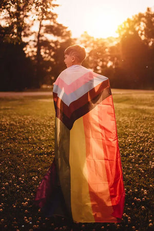 A person standing in the light of a sunset wearing the Progress Pride flag like a cape.