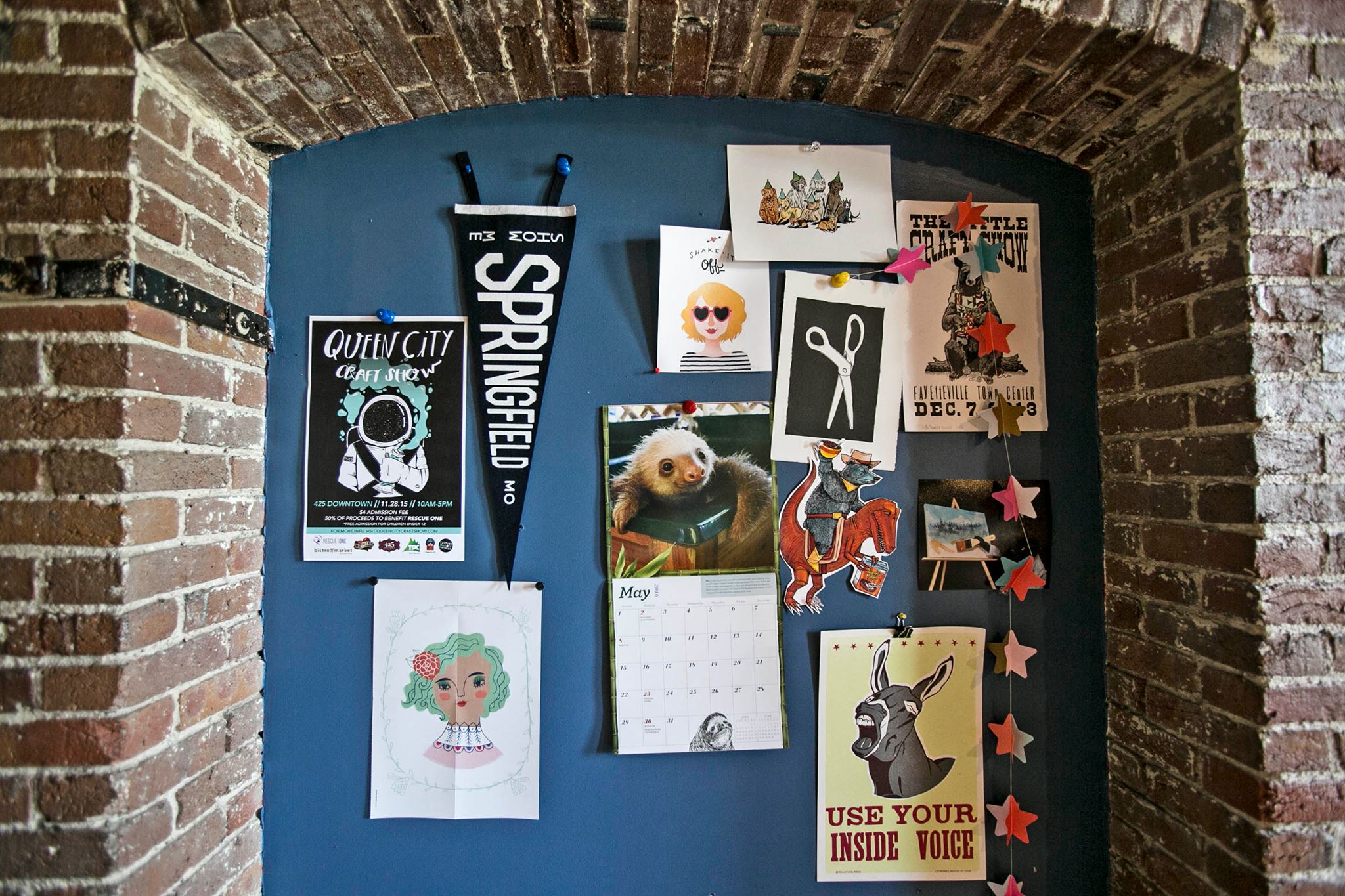 Blue bulletin board with various magnets and a calendar against a brick wall.