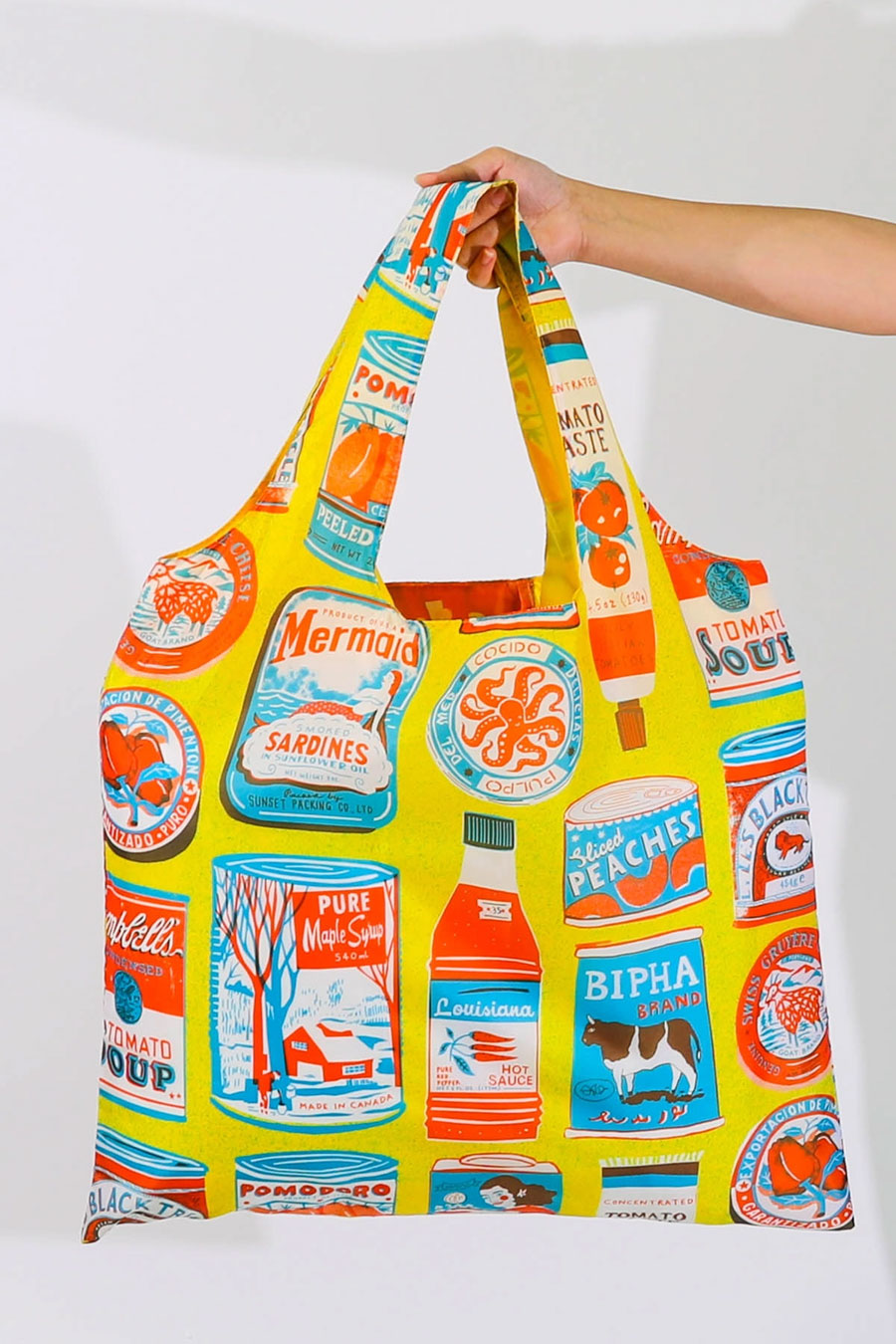 Yellow reusable tote bag covered in all over print of illustrated food products in white, red, and blue.