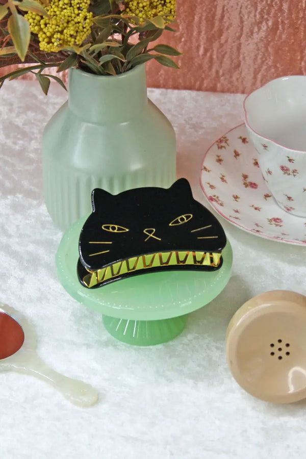 Black kitten face claw hairclip on a green glass pedestal, next to a floral tea cup, a vase of flowers, a vanity mirror, and a phone receiver.