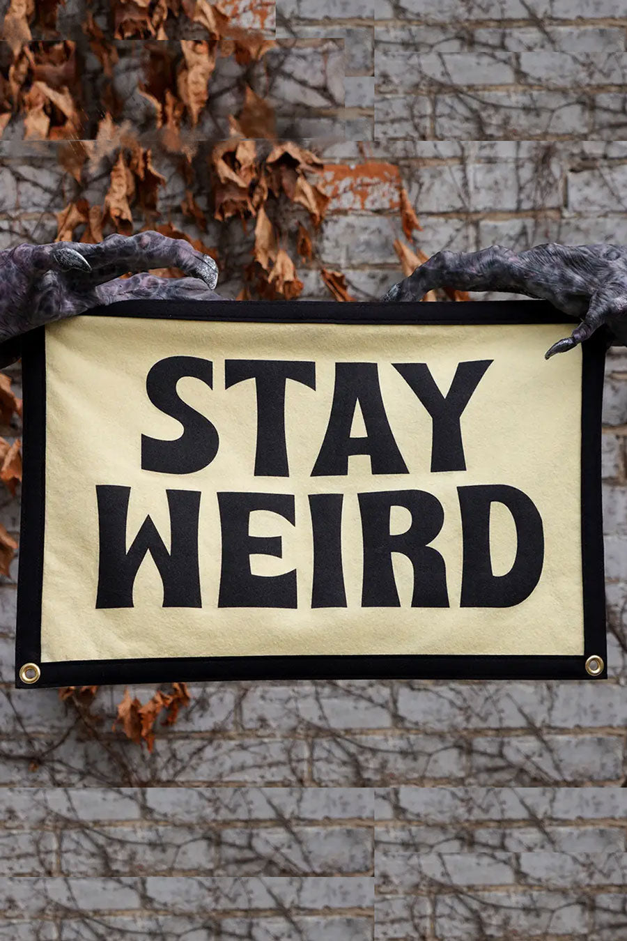 Two monster claws holding Ivory camp flag with black border and four grommets, one on each corner. The flag says Stay Weird in bold black lettering against a gray brick wall.