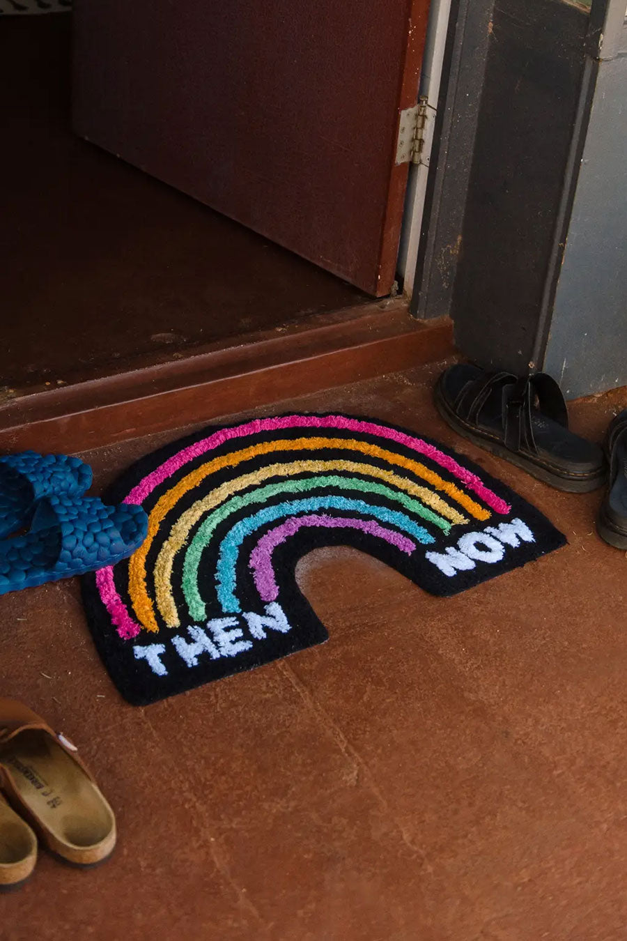Black rainbow shaped rug featuring rainbow color striped arches. On the left side under the stripes the rug says Then and on the right at the end of the stripes the rug says Now. This rug is at the doorstep of a house surrounded by sandals.