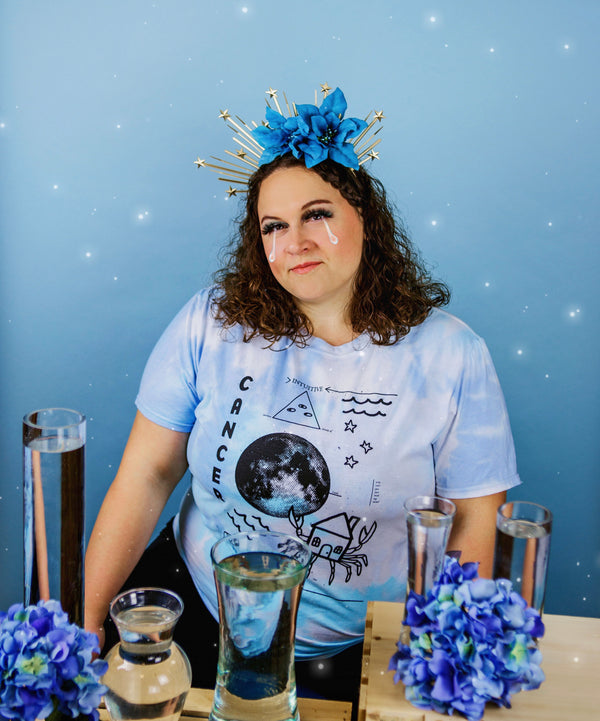 A woman with dark curly hair wearing the blue tie-dyed tee depicting the astrological symbols for "Cancer." She is sitting at a table with vases full of water, wearing a flower crown.