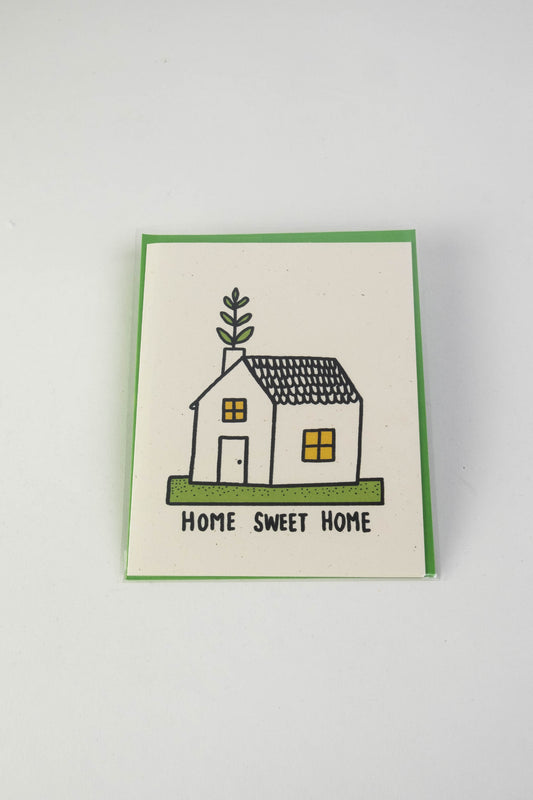 A white greeting card with the illustration of a small house with green foliage coming out of the chimney. The text along the bottom reads "Home sweet home."