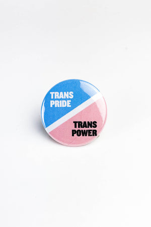 Round pinback button on white backgorund. The button is half blue and half pink with a white diagonal stripe to that separates the two colors. In the top blue section it says Trans Pride in white. In the pink portion it says Trans Power in black text.