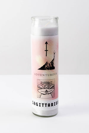 A yellow and pink decal is affixed to a tall glass votive candle. The candle is sitting on a flat white surface. The decal has illustrations symbols attributed to the zodiac sign Sagittarius and features text that reads "Sagittarius," "Adventurous."
