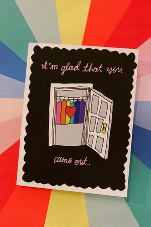 A photo of a greeting card against a rainbow starbust background that reads "I'm so glad you came out" and an illustration of an open closet door with rainbow clothing inside. The background of the photo has diagonal rainbow panels.