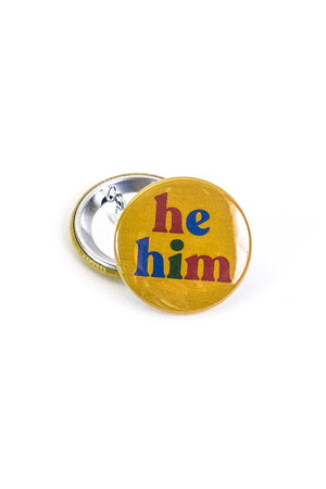 Yellow button laying on top of a turned over pinback button on a white background. The button says "He Him" in alternating letter colors of blue red and green.