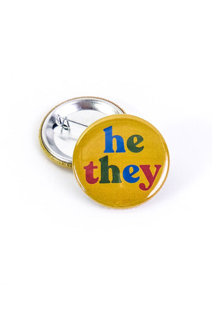 Yellow button laying on top of a turned over pinback button on a white background. The button says "He They" in alternating letter colors of blue red and green.