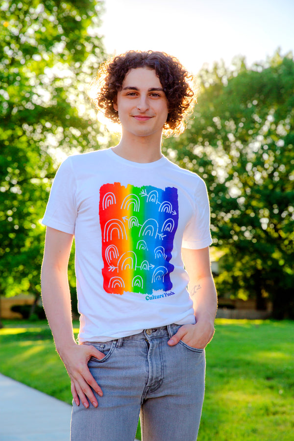 A person with dark curly hair is standing on the sidewalk in front of a lush green yard and big trees. They are wearing a white t-shirt with a vertical rainbow flag and small white rainbows overlayed. 