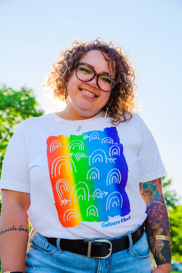 A woman with dark curly hair is standing in the sun in front of green trees. She is wearing jeans, a black belt, and a tucked in white t-shirt with a rectangular vertical rainbow pride flag and white illustrated rainbows overlaying the flag. 