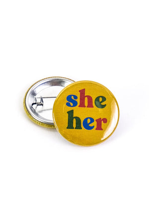 Yellow button laying on top of a turned over pinback button on a white background. The button says She Her in alternating letter colors of blue red and green.