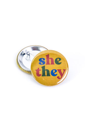 Yellow button laying on top of a turned over pinback button on a white background. The button says "She They" in alternating letter colors of blue red and green.