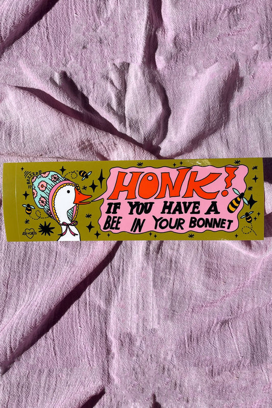 An olive green bumper sticker depicting a white goose wearing a bonnet. The pink text bubble reads "Honk if you have a bee in your bonnet" on a purple linen backdrop.
