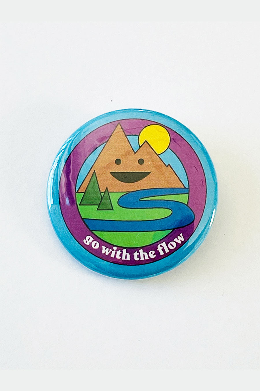 A pinback button with a blue background and an illustration of a smiling mountain range, a river, and trees. The text reads "go with the flow."