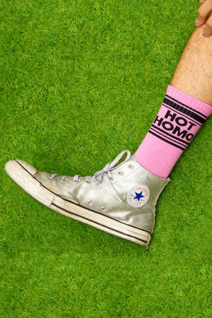 Photograph of someones leg wearing silver shoes and midcalf pink crew socks that say "Hot Homo" across the top. The socks have a black stripe above and below the text. Grass background.