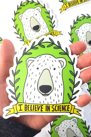 A hand is holding a die-cut sticker of a white polar bear surrounded by green laurel. The bottom has a yellow banner that reads "I believe in science" in black lettering. In the background are several more of the sticker laying on a flat surface.