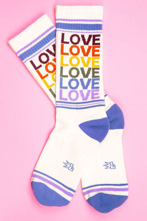 Vintage white mid-calf crew socks. The socks have a light blue heel and toe with a thin blue and purple stripe across the toe and across the top of the socks. The socks say Love in a repeating pattern in vintage looking colors of Burgundy, orange, yellow, sage green, light blue, and lilac purple. Pink background.