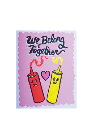A pink greeting card with an illustrated purple scalloped border says "We Belong Together" in black text. Below are illustrations of a bottle of ketchup and a bottle of mustard with a pink heart in between them.