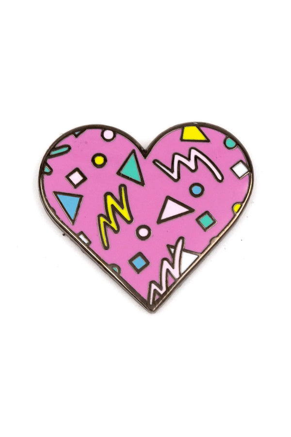 Enamel pin of a pink heart with teal, white, and yellow confetti and zig zag patterns. White background.