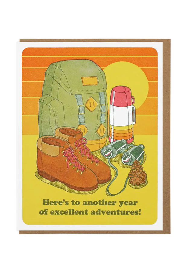 White greeting card featuring illustration of hiking boots, backpack ,striped thermos, binoculars, and a pinecone sitting in front of a sun and a striped sky. The card says Here's to another year of excellent adventures. White background.