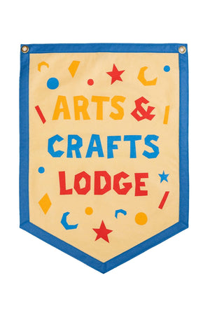Beige Camp flag with a blue border. The Flag says Arts & Crafts Lodge in yellow, blue, and red text surrounded y different shapes in the same colors.