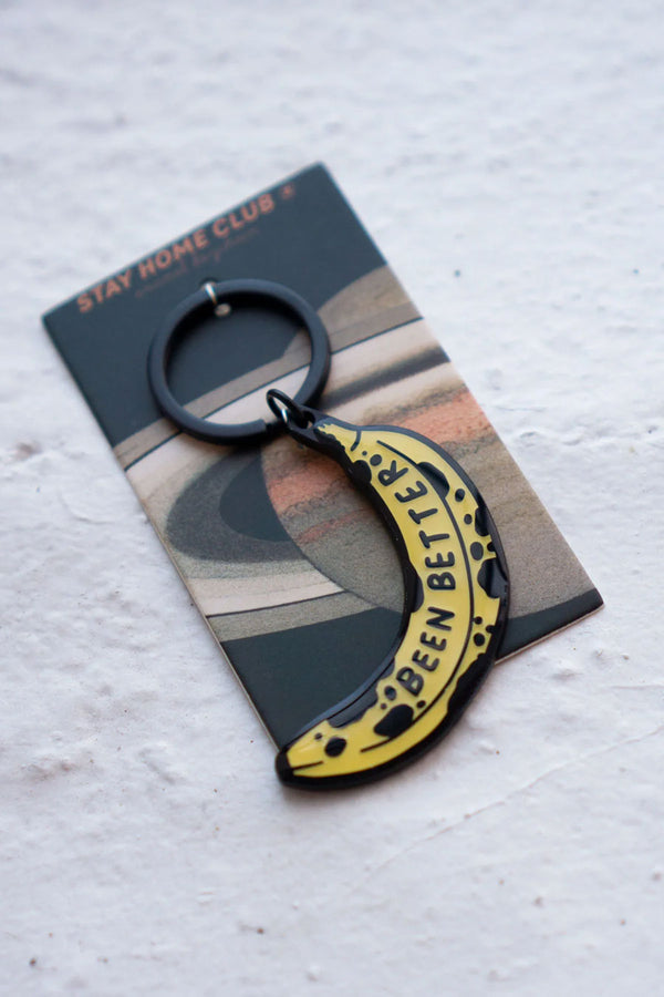 Enamel keychain with black hardware of a banana with black spots. The banana says Been Better in black text. The keychain is attached to a backing board that features Saturn and says Stay Home Club at the top.