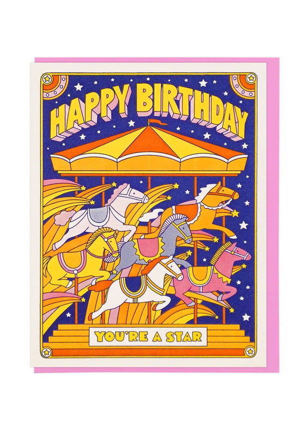 Greeting card of a carousel of horses. Above the carousel the card says Happy Birthday in Yellow block letters. On the base of the carousel it says your a star. There are yellow, pink, and orange star bursts among the horses against a blue sky. Pink Envelope. White background.