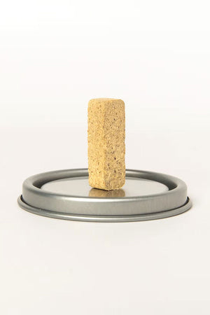Photo of a small incense brick on top of a metal container lid. White background.