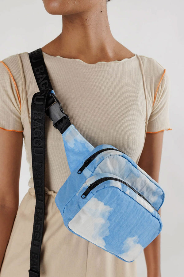 Person wearing a nylon fanny pack across their body. The fanny pack features black straps and realistic clouds against a blue sky printed all over.  White background.