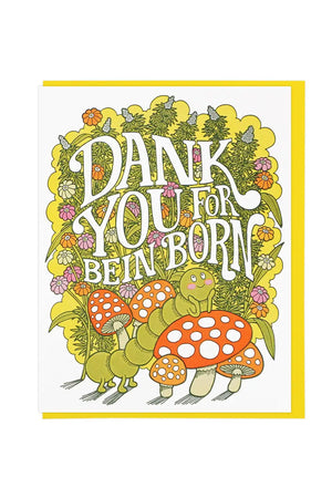 White greeting card of a marijuana plant with pink, yellow, and pink flowers in it. In front of the plant is red mushrooms with a green caterpillar with a smiley face. The card says Dank You for Bein' Born in white lettering on top of the plant. Yellow envelope. White background.