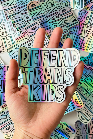 A photo of a hand holding a holographic die cut sticker that says Defend Trans Kids in block lettering. This sticker is being held over multiple of these stickers on a white background.