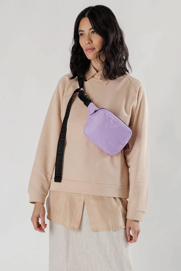 Person standing in front of a white backdrop wearing a khaki sweater and skirt with a lilac fanny pack across their chest. The fanny pack has black nylon strap that says Baggu in a repeating pattern.