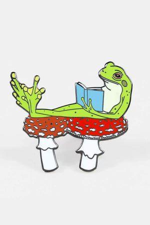Enamel pin of a Green Frog laying on two mushrooms reading a book