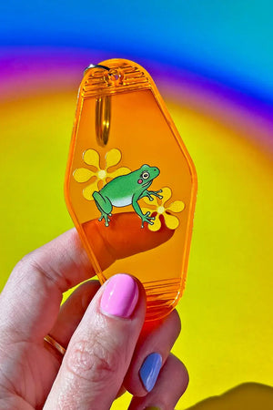 Person holding an orange transparent vintage motel shaped keychain. The keychain features a frog with yellow flowers. Rainbow background.