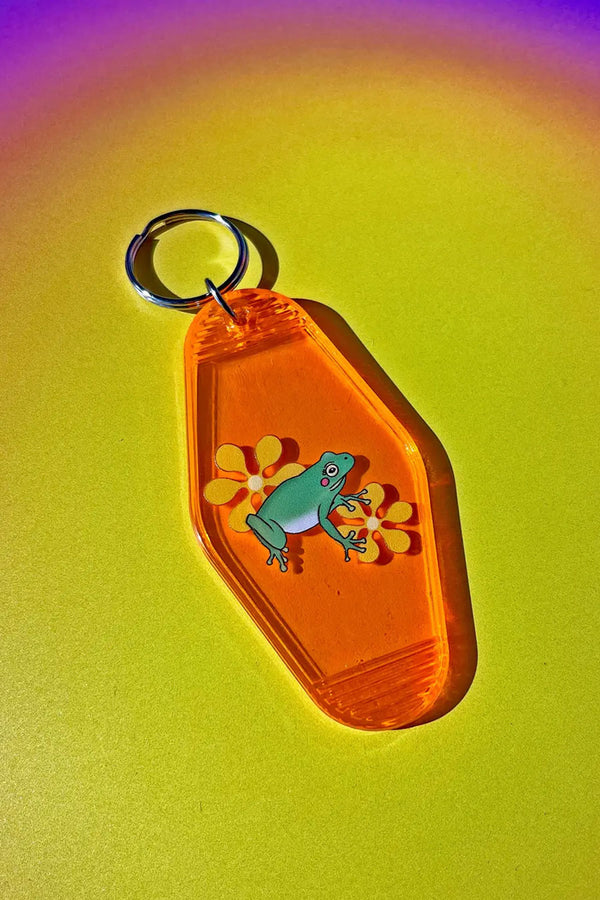 An orange transparent vintage motel shaped keychain. The keychain features a frog with yellow flowers. Rainbow background.
