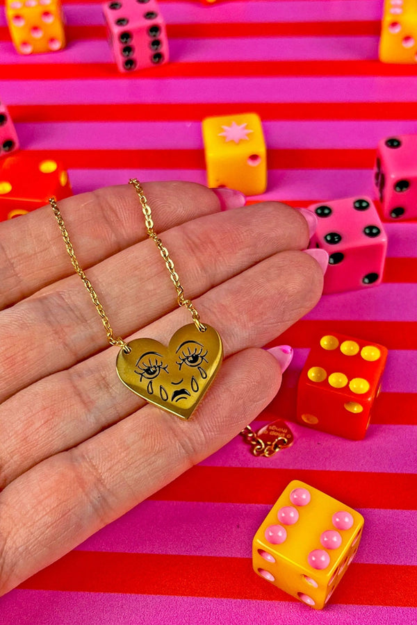 Someone holding a Gold heart necklace with gold chain. The heart features a crying face. Red and Pink striped background.