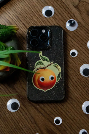 Vinyl sticker of a peach with googly eyes on the back of a cell phone. The phone is surrounded by random googly eyes laid on a table.