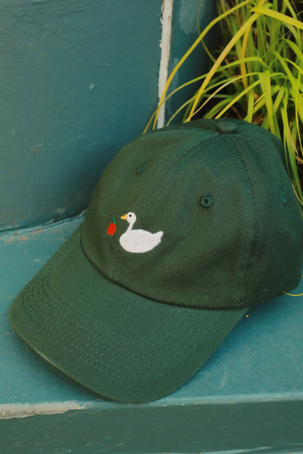 Green dad hat featuring embroidered design of a white goose and a red apple. Hat is on green steps next to a plant.