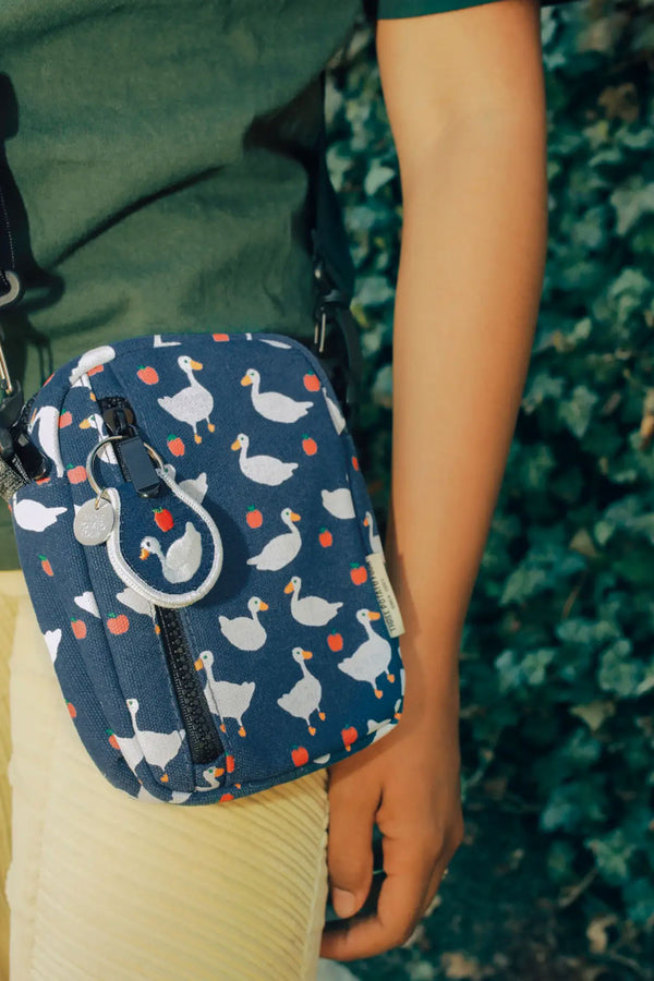 A person wearing a sling bag across their body. The bag features a pattern of white geese and apples all over. 