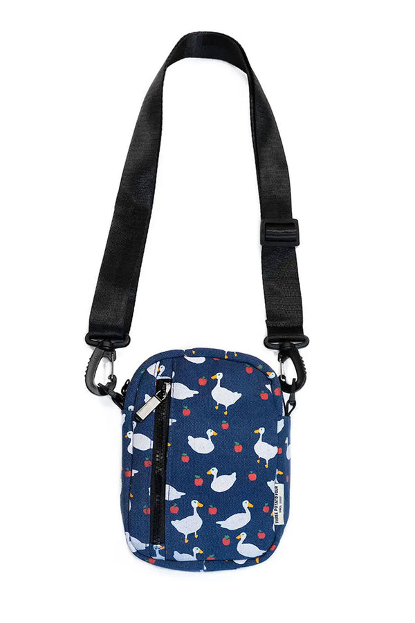 Navy sling bag with black straps featuring an all over pattern of white geese and apples. White background.