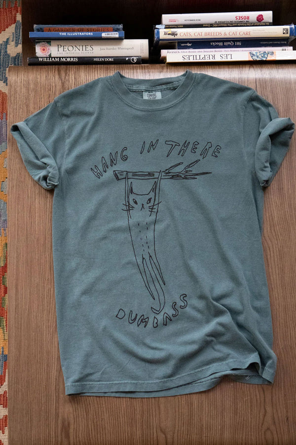 A green tshirt that features an illustration of a cat hanging from a tree branch. The shirt says Hang in there Dumbass. Wooden table background.
