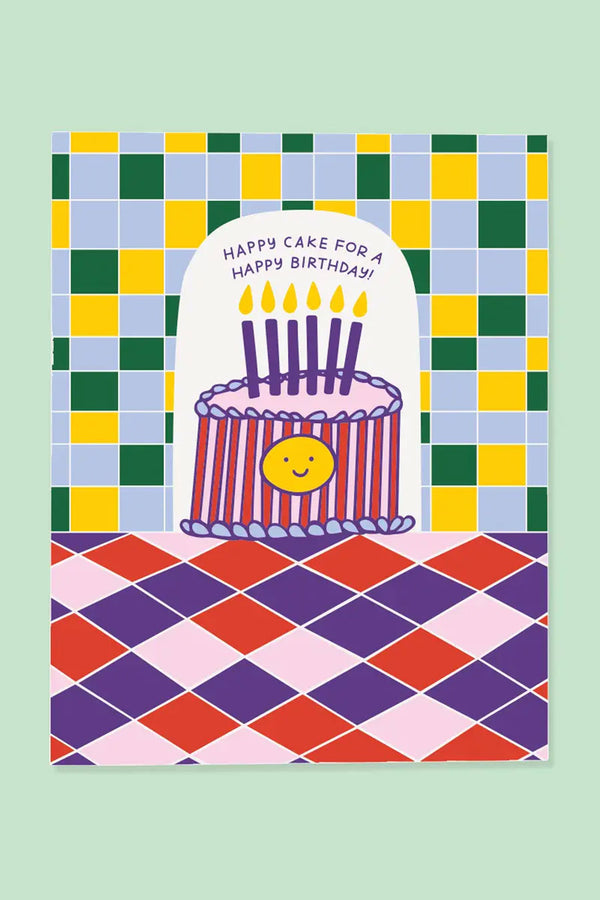 Birthday card featuring a cake with purple candles and red and pink stripes with a yellow happy face on the front of it. Card says Happy Cake for a Happy Birthday! Back ground is blue, green, and yellow checkered pattern. The bottom half is a diamond pattern of pink, purple, and red.