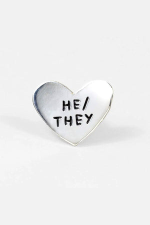 Silver heart shape enamel pin. The pin says He/They in black text. White background.