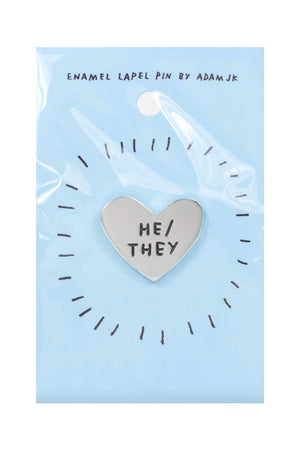 Silver heart shape enamel pin. The pin says He/They in black text. The pin is on a blue backing board. White background.