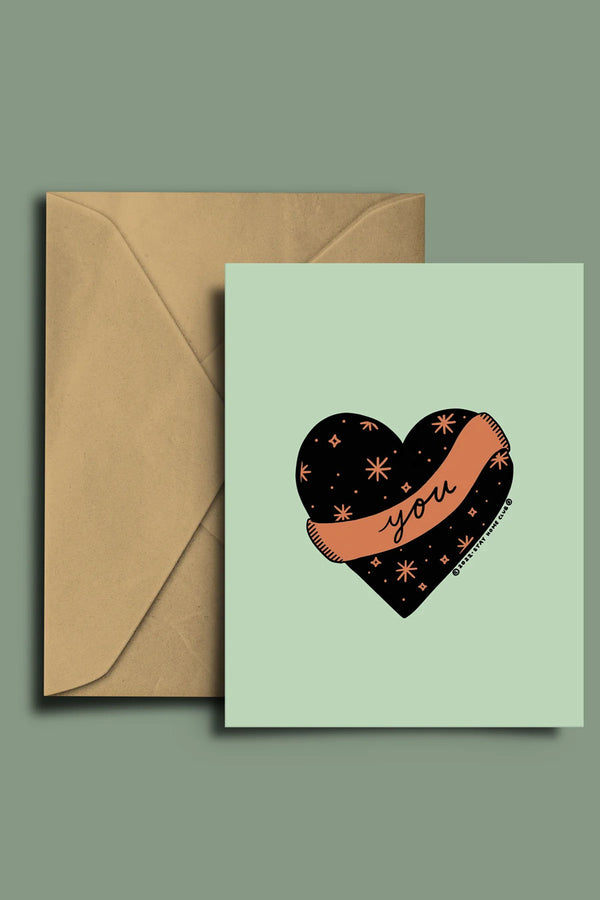 Light green card with kraft brown envelope against a sage green background. The card has a black heart with a pink banner around it. The heart has pink stars all over it and the banner says You in cursive.