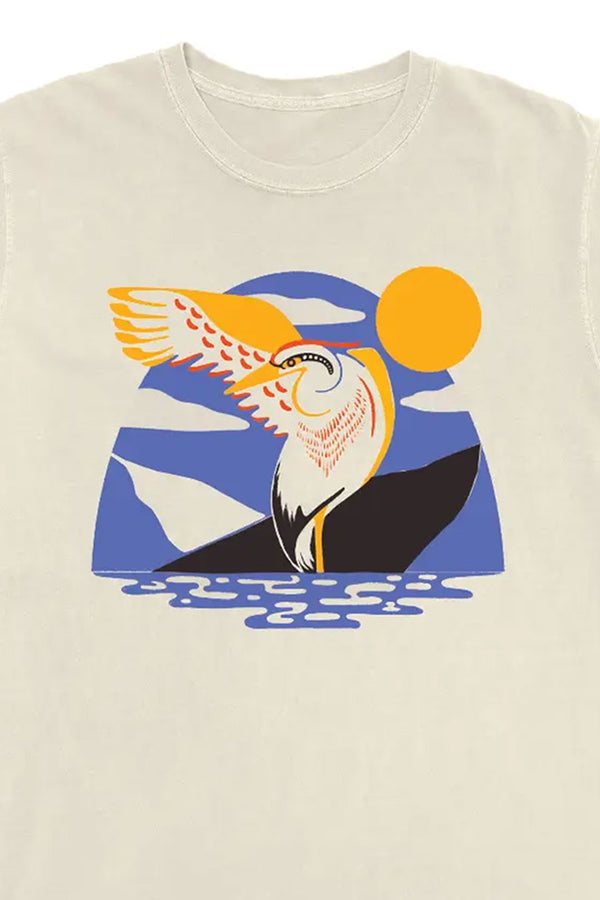 Close photograph of a Natural color tshirt against a white background. The shirt features a modern illustration of a heron standing in the water with one wing outstretched. Behind the heron is a blue arched sky with a yellow sun in the sky. The design is made of a limited color palette of yellow, red, blue, and black.