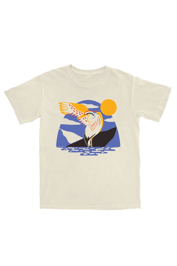 Natural color tshirt against a white background. The shirt features a modern illustration of a heron standing in the water with one wing outstretched. Behind the heron is a blue arched sky with a yellow sun in the sky. The design is made of a limited color palette of yellow, red, blue, and black. 