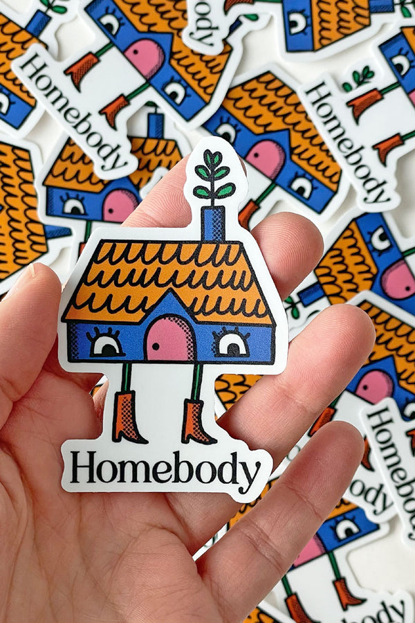 A person holding a Die cut sticker of a blue house with a yellow roof and a chimney. The house has a pair of eyes for windows, a pink door, and a plant coming out of the chimney. The house also has a pair of legs wearing red boots standing over text that says Homebody. This sticker is being held over multiple stickers on a white background.