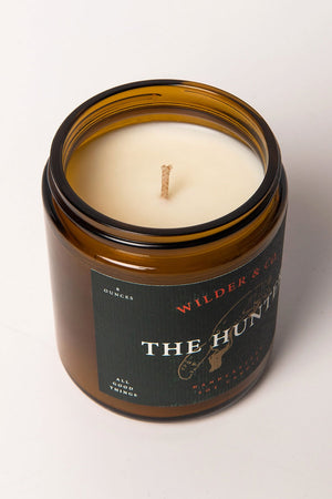 Candle in amber glass jar with forest green label. Label features an illustrated jumping fox and says Wilder & CO, The Hunter.